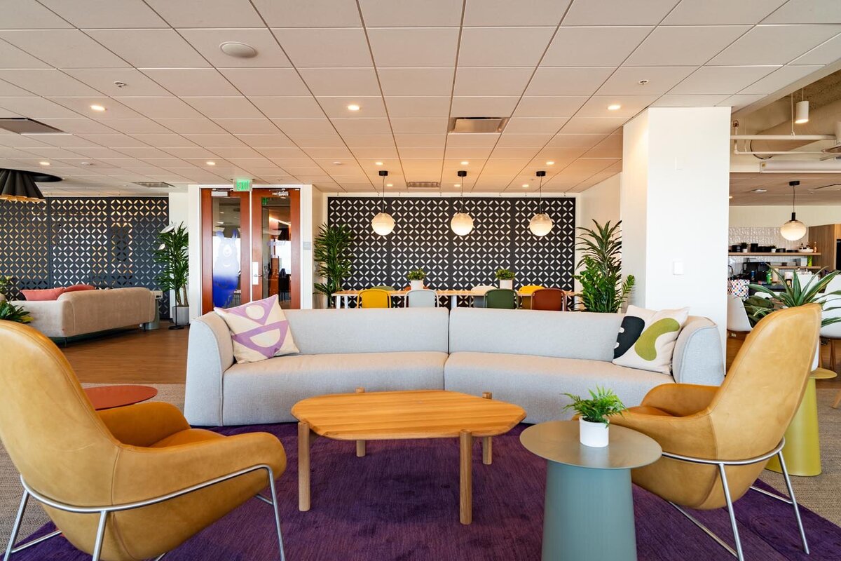 Colorful office lounge with grey sofa, tan leather chairs, purple rug, and geometric patterned feature wall.