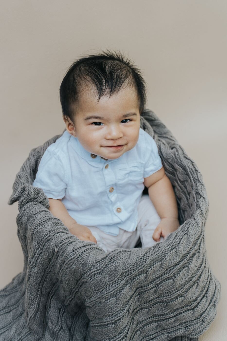 Baby boy sitting upright inside a chunky gray knitted blanket, wearing a light blue shirt and smiling sweetly.