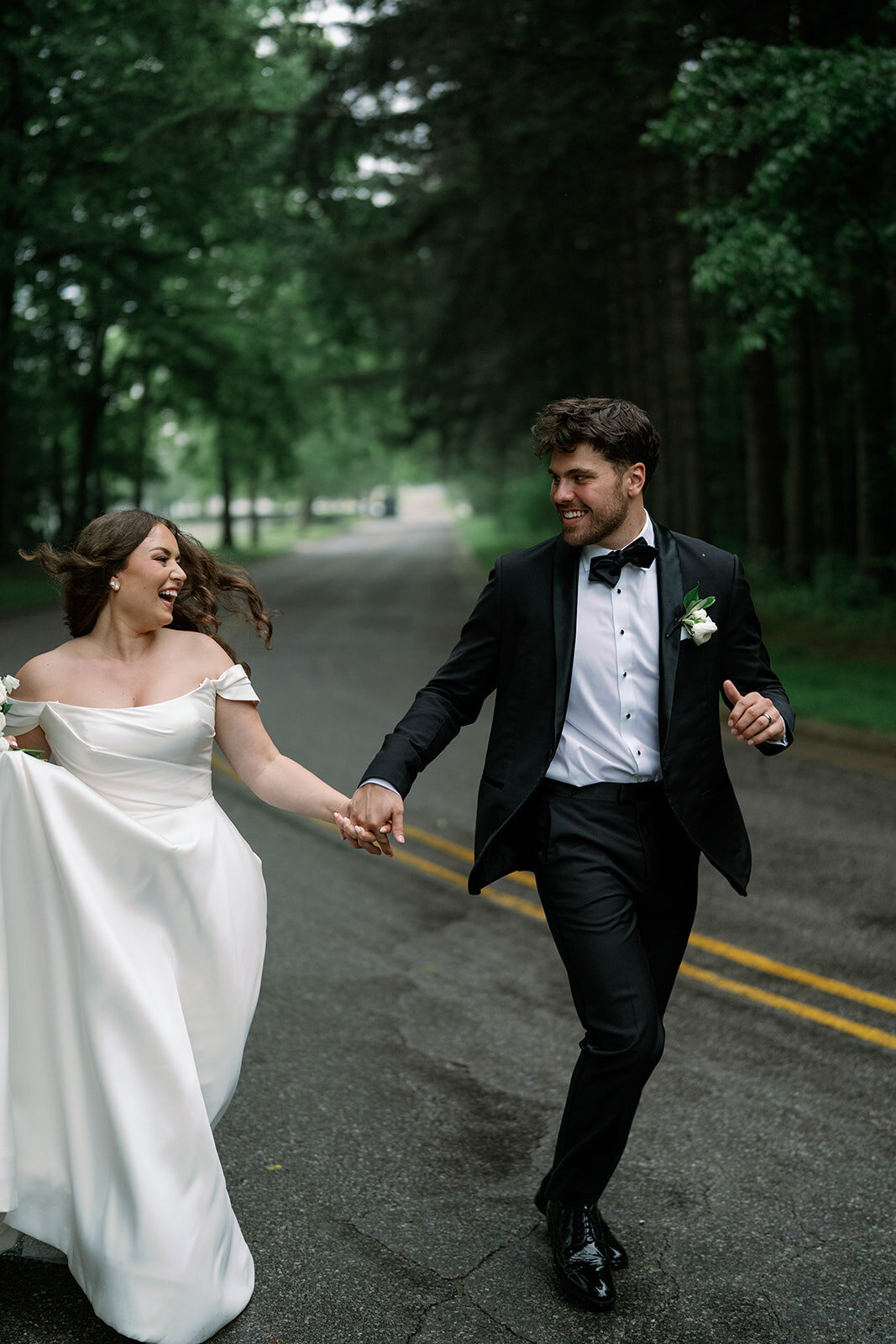 Bride and groom joyfully running down a tree-lined road near The Morris Estate during their wedding portraits.
