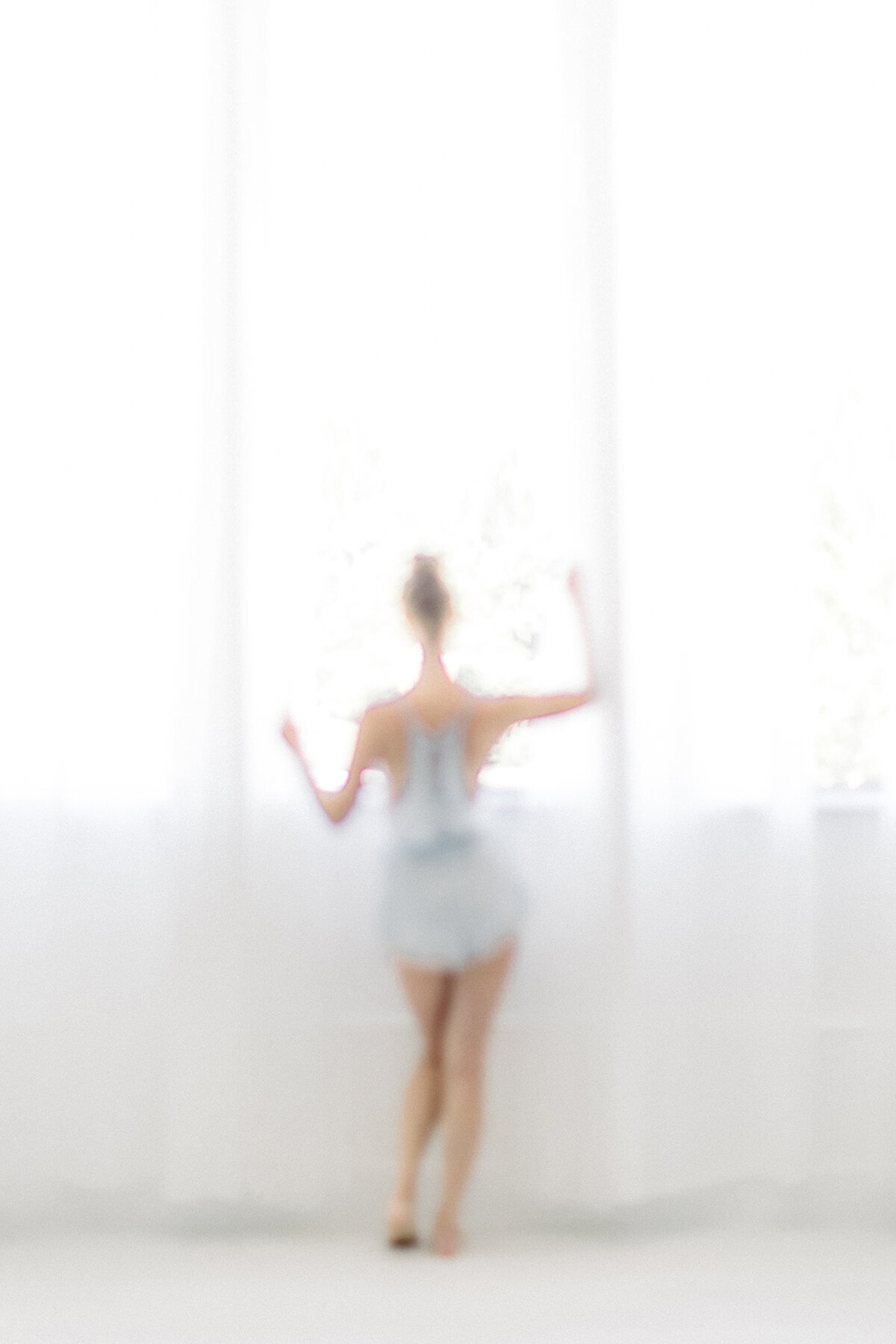 A light filled out of focus photo of a woman dressed in a lace romper standing in sheer drapes looking out of the window of a Dallas TX photography studio.