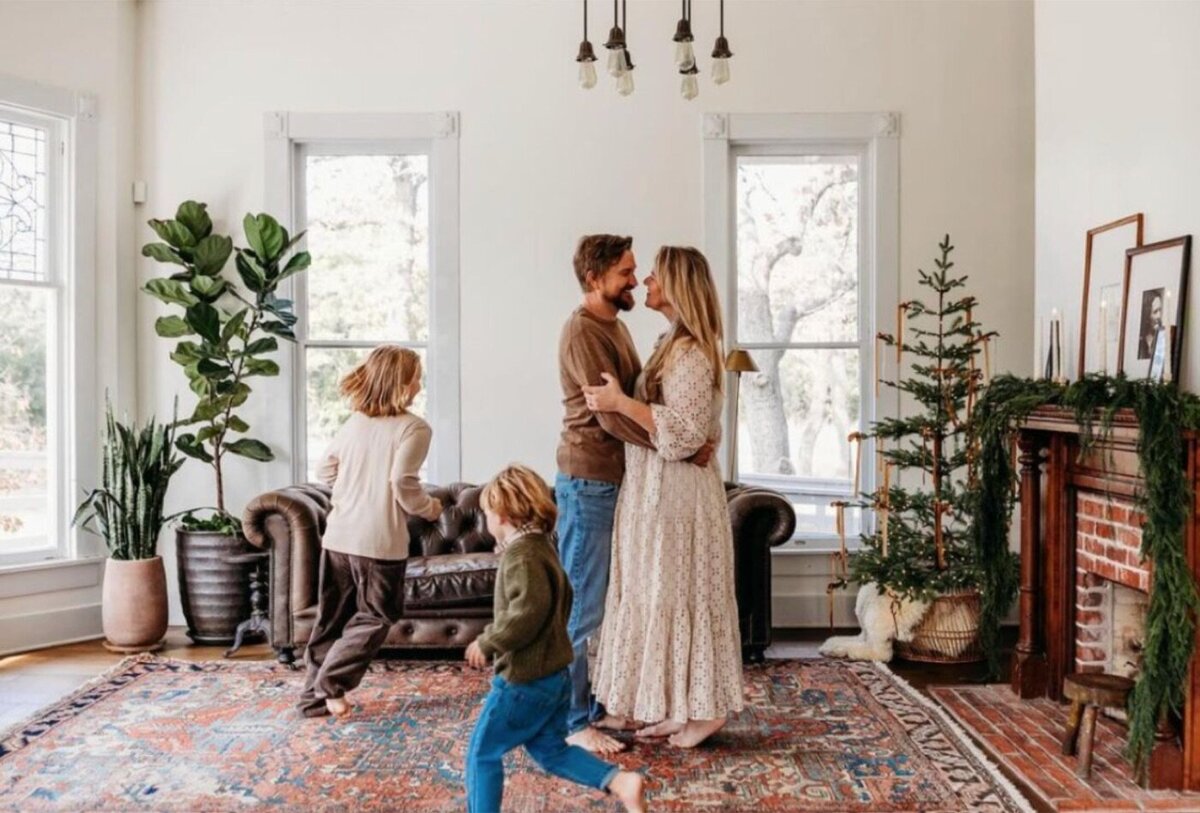Family dancing in the mansion living room beside a brick fireplace with holiday greenery
