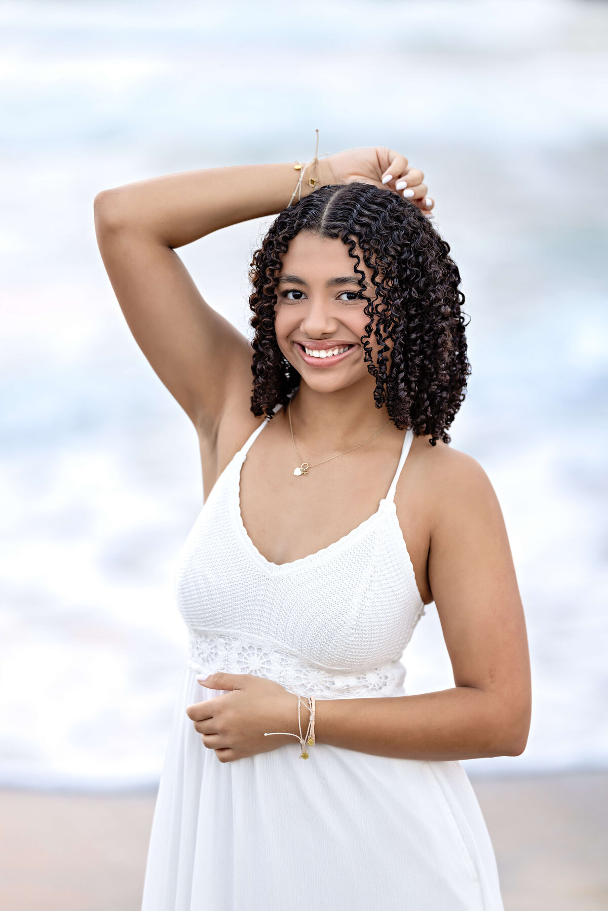 Orlando senior session close-up of a girl with curls and a white dress, one arm raised, soft smile, glowing against the pastel sky.