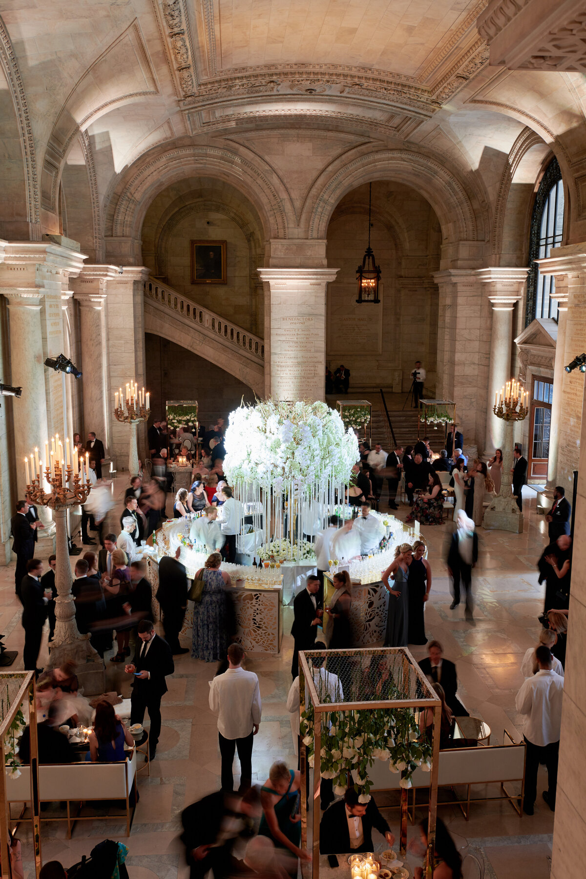The photo was taken at The New York Public Library in New York City by photographer Christian Oth. The grand, spacious hall with arched ceilings and columns exudes elegance as it hosts an elegant event. Guests in formal attire mingle around a large floral centerpiece, adding to the luxurious atmosphere. In the background, a majestic staircase enhances the grandeur of the setting, while chandeliers cast a warm glow over the sophisticated ambiance. Additionally, East West Films provided videography services for this event.