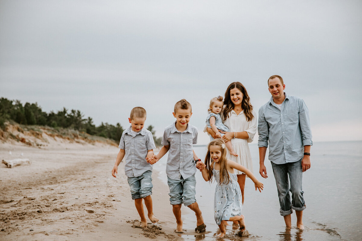 Mom, Dad, and four young children are walking along the shoreline at Grand Bend beach for a family photoshoot. Mom and Dad are smiling at the camera, mom holding the youngest daughter.  The three older children are holding hands in front of of them and running in the water.