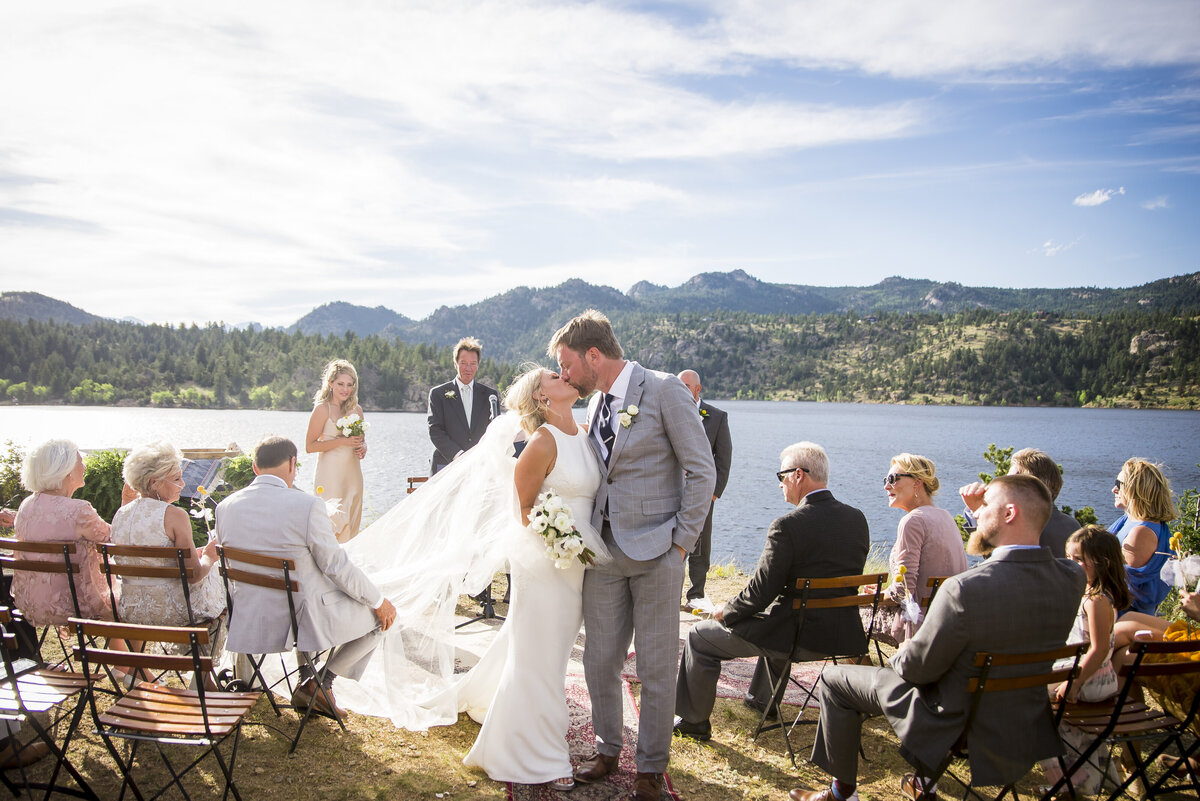 The bride and groom share their first kiss as husband and wife in front of a lake and mountain background.