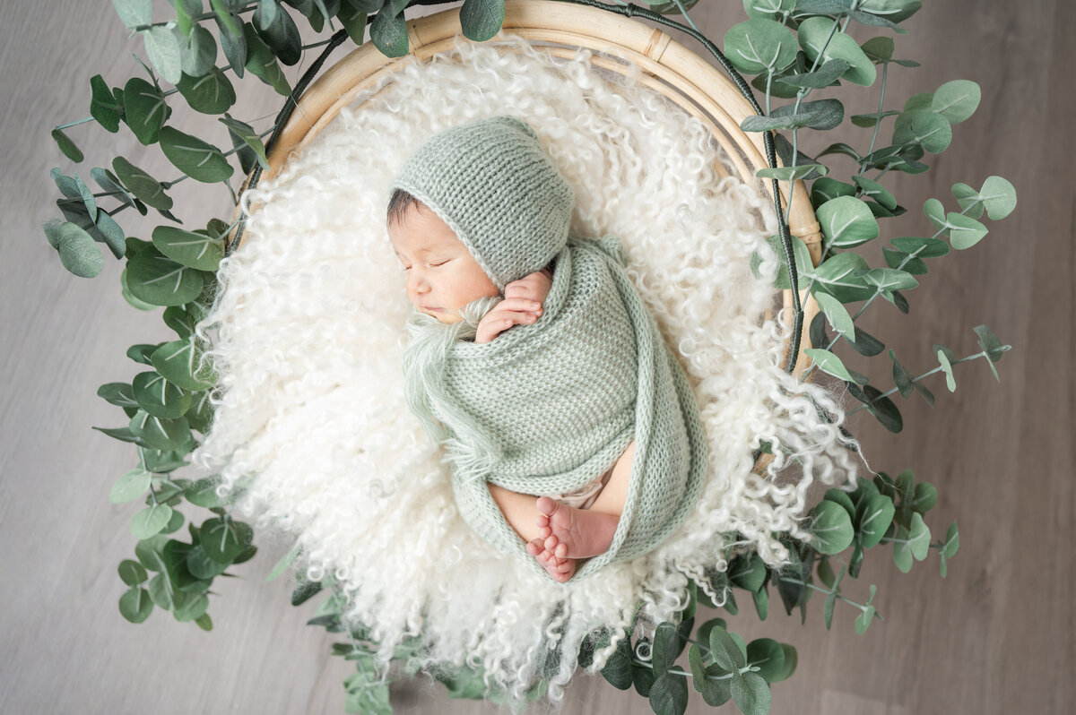 Newborn baby in sage green wrap and bonnet, resting on a cream flokati with eucalyptus accents.