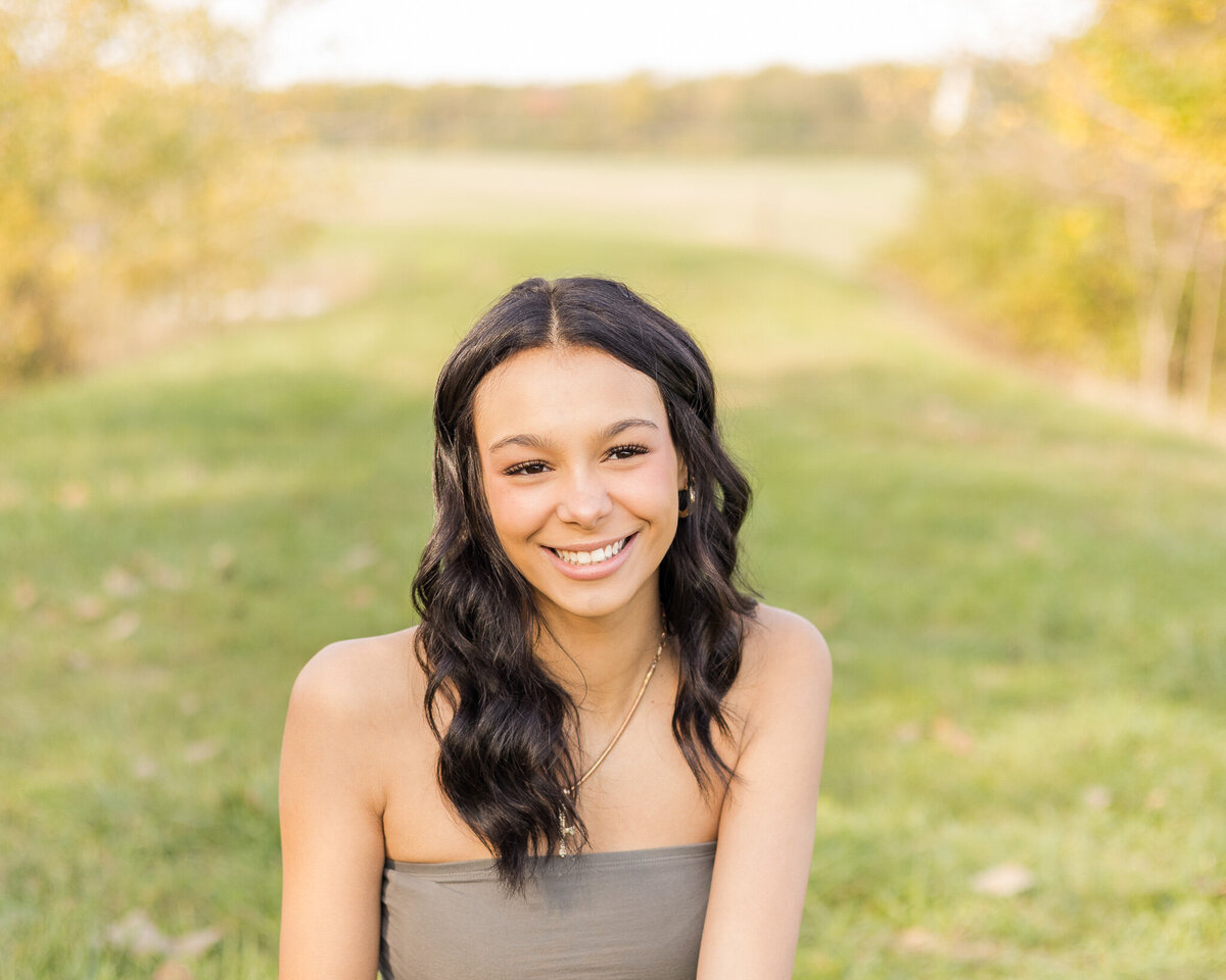 A high school senior female with long black hair, sitting in grass with trees in the background, wearing a sleeveless green shirt, smiling away from the camera