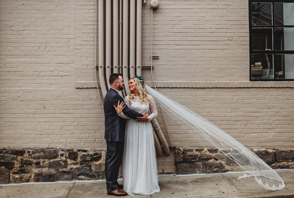 Maryland wedding photographer photographs outdoor wedding pictures in downtown while the brides veil blows in the wind as she looks to her room and he holds her waist