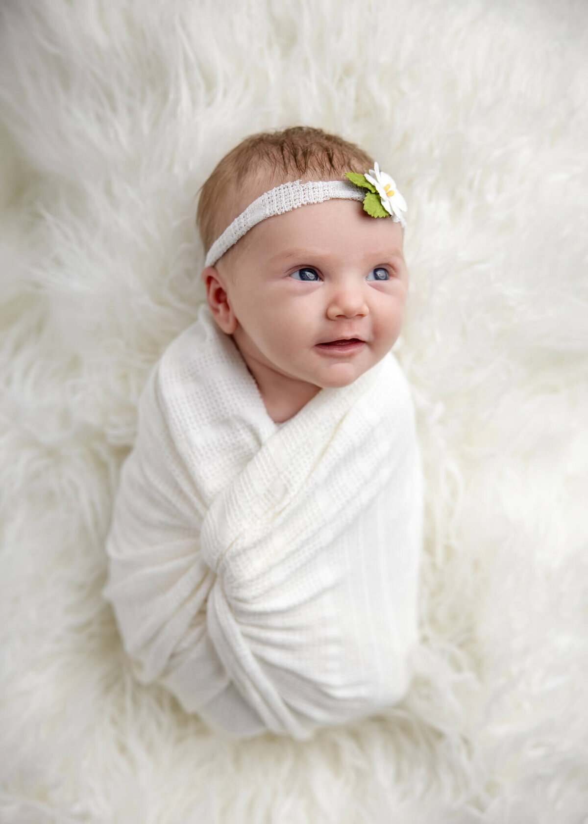 smiling newborn baby wrapped in white fabric wearing a white floral headband laying on a white shaggy rug
