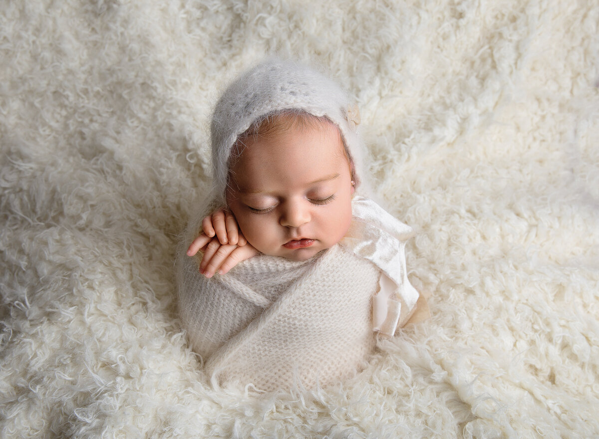 Baby girl is swaddled in cream and placed atop of a fuzzy cream flokati. Baby is wearing a matching bonnet. Captured by top Brooklyn newborn photographer Chaya Bornstein
