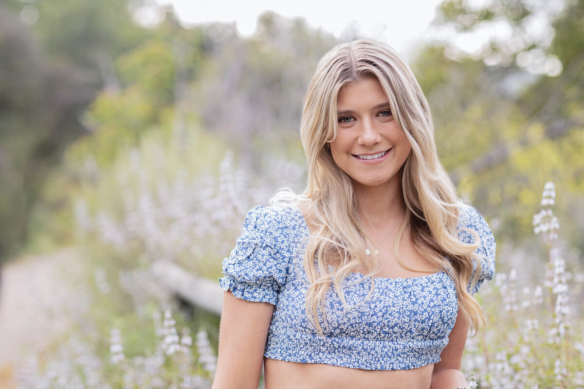 A close-up of a young woman with long, blonde hair standing in a field of flowers and greenery, wearing a blue and white floral cropped top and smiling at the camera in San Juan Capistrano.