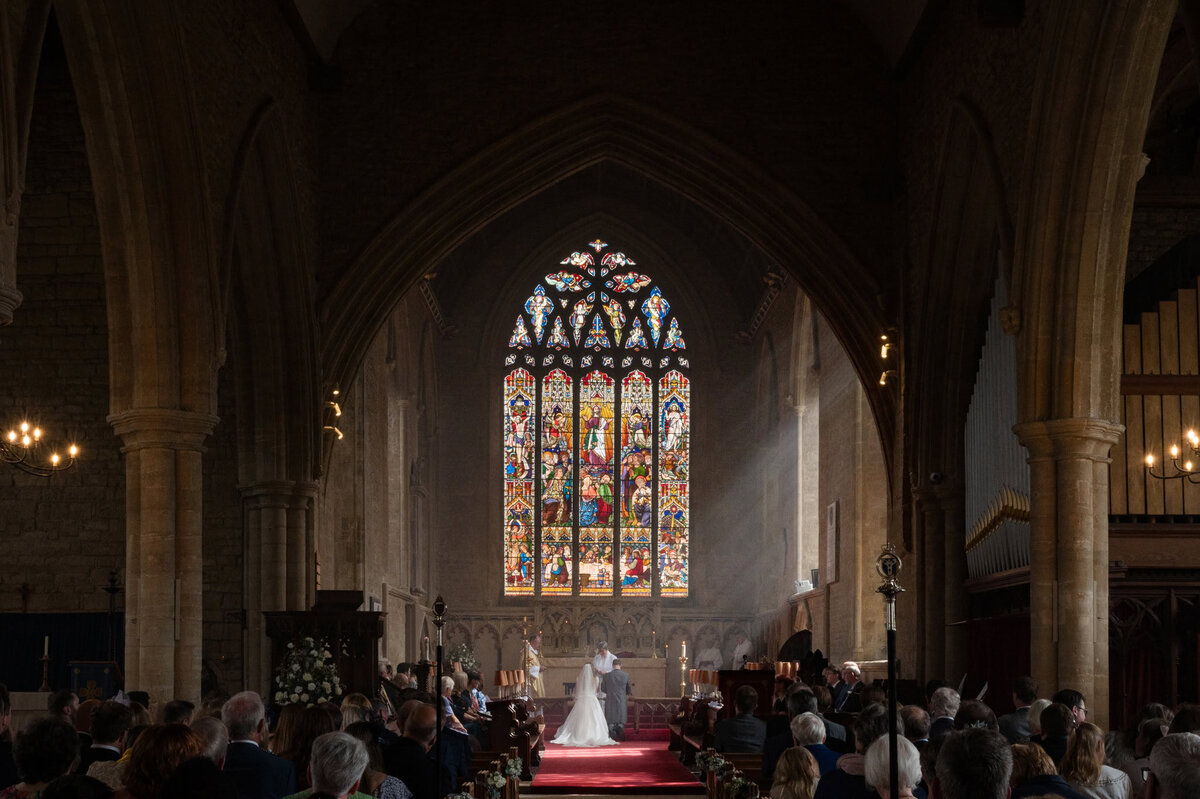 Sunlight streaming in through church stained glass windows as bride and groom kneel for prayers