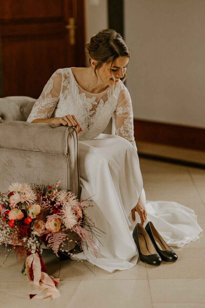 Mariée assise dans un fauteuil, bouquet à ses pieds, s'apprêtant à enfiler ses escarpins noirs, posant pour Laura lors d'un workshop photographie de mariage.