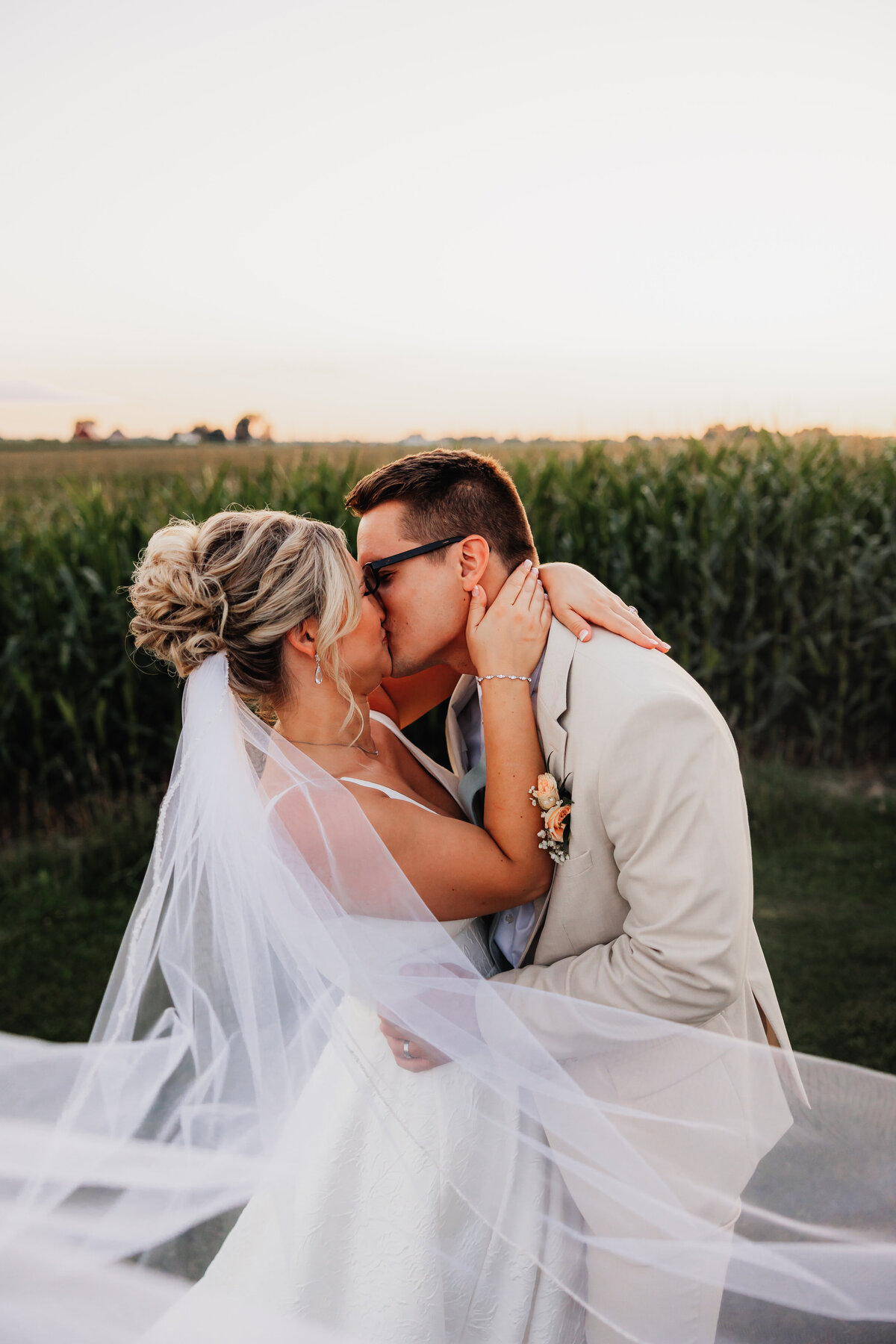 A Bride and groom share a passionate kiss in front of a corn field.
