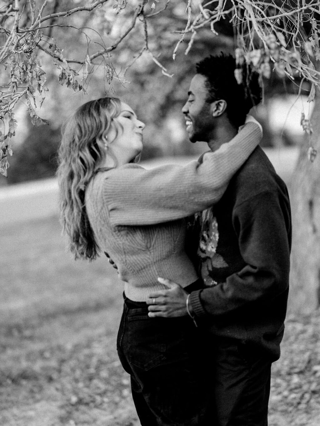 A couple hugs under a tree in Cheesman Park. They are looking at each other and laughing.