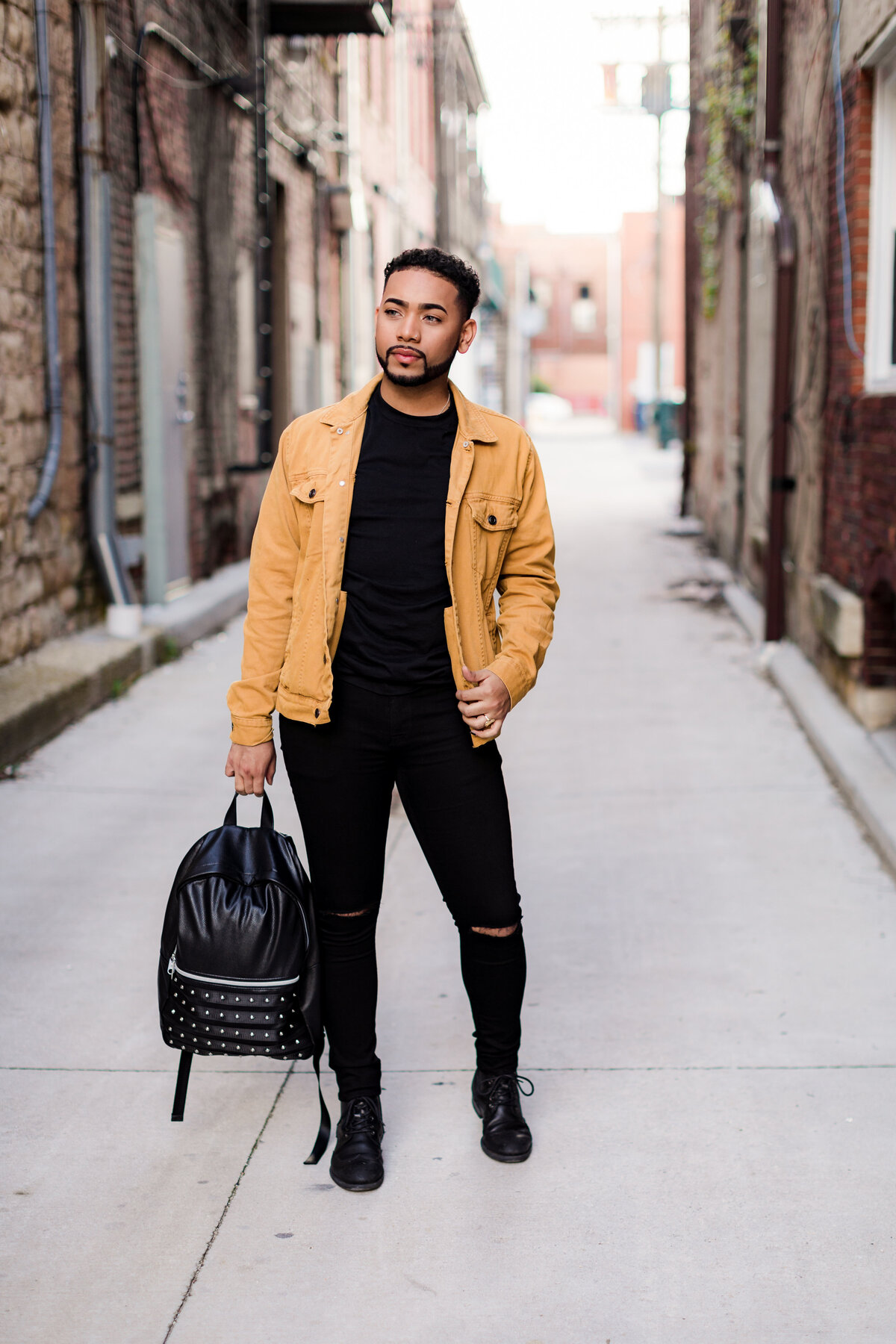 A young man wear a black outfit with a yellow jacket while holding a backpack by his side in an alleyway.