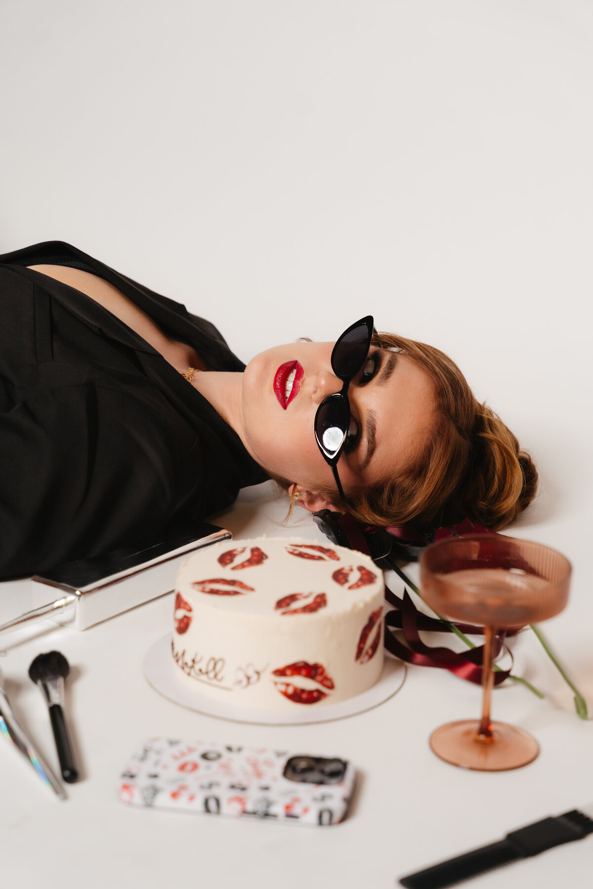 Makall lying on her side in black blazer and red pants, wearing cat-eye sunglasses — bridal artist portrait