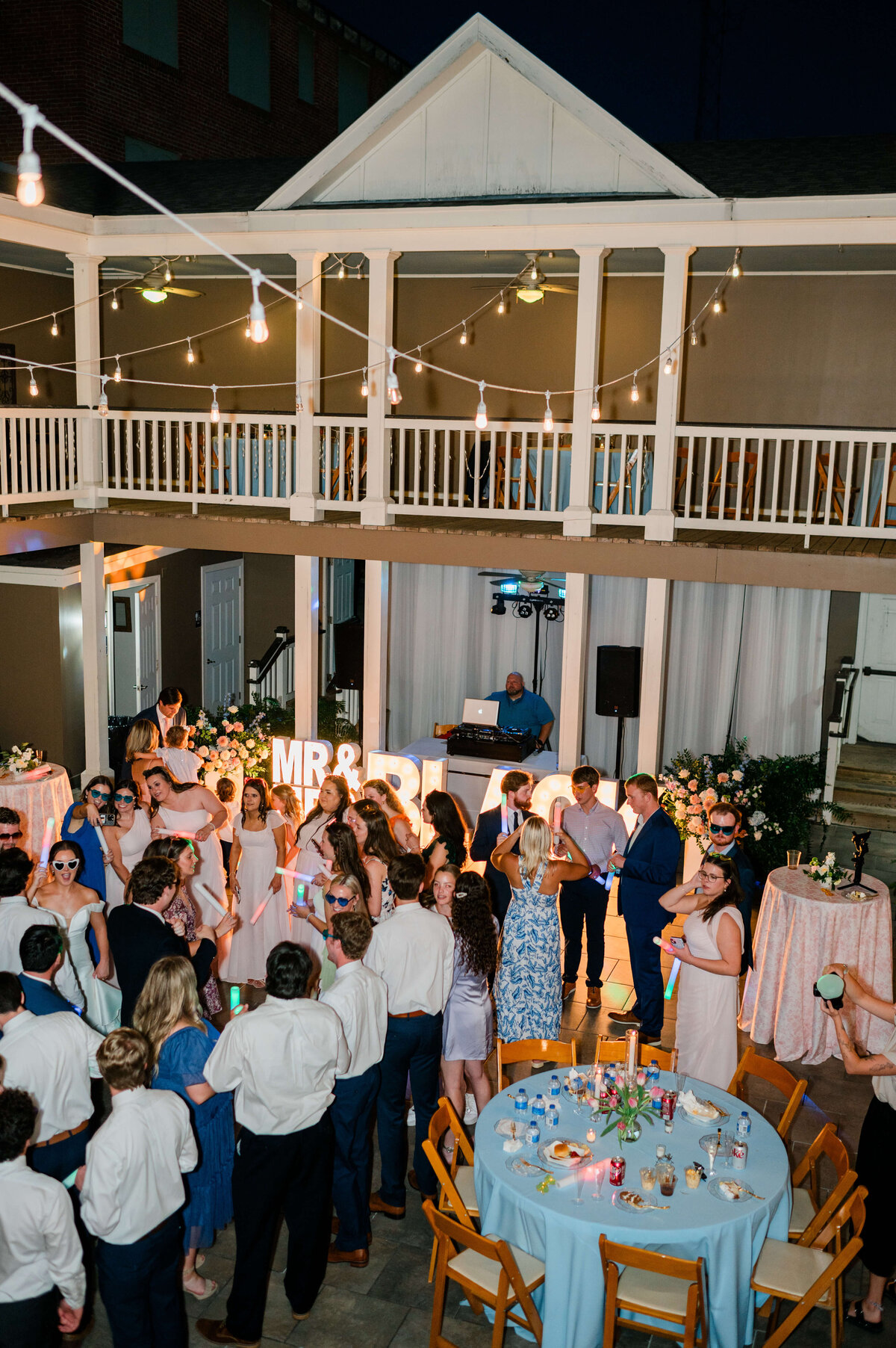 A lively outdoor wedding reception at night, with guests dancing under string lights. Tables are decorated with flowers and candles.
