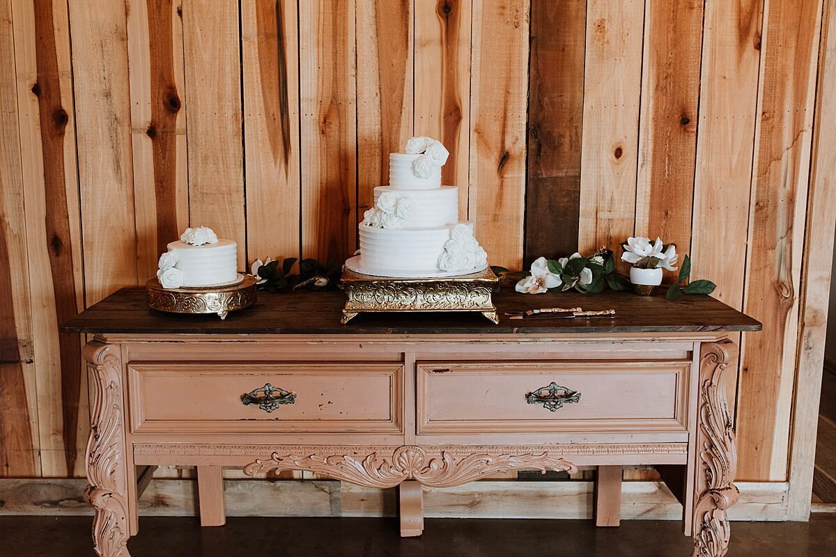 Decorative brown dresser with two drawers has a small white grooms cake on a round silver cake stand and a larger three tier white wedding cake on a square silver stand with white flowers against a wooden wall at Steel Magnolia Barn.