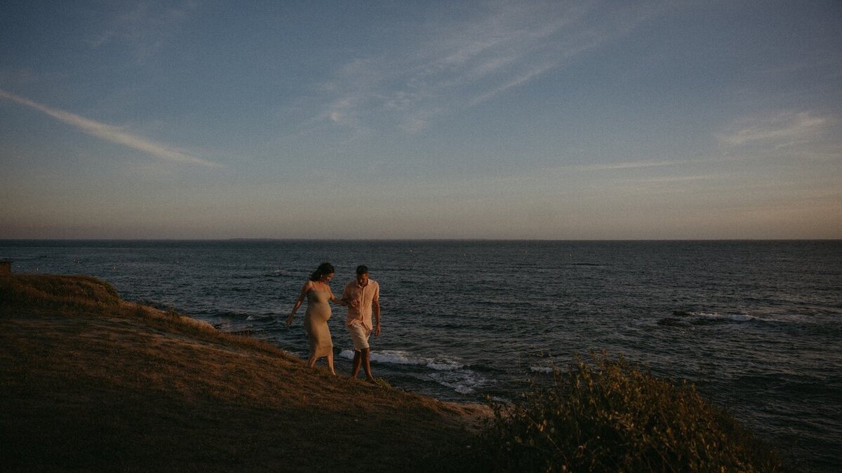 Le couple marche au loin sur une falaise, face à l’océan et au coucher du soleil. Shooting grossesse par Kamescop, vidéaste et photographe en Bretagne.