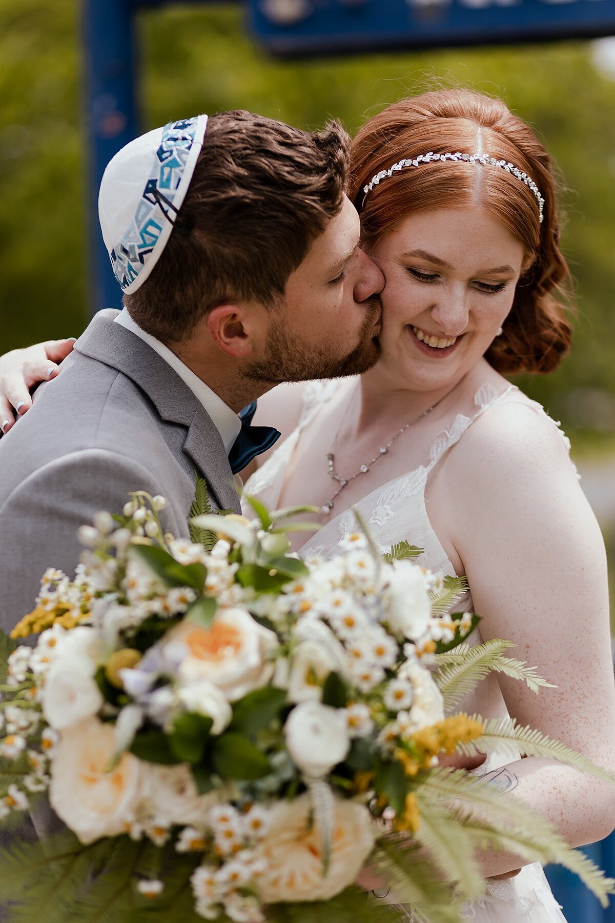A redheaded bride wearing a v-necked white floral wedding dress holding a large bouquet of white and yellow wildflowers and fern as she laughs. The Jewish groom wearing a blue and white kippot and grey suit leans in to kiss the bride's cheek at their Jewish Wedding in Nashville, TN at Loveless Barn