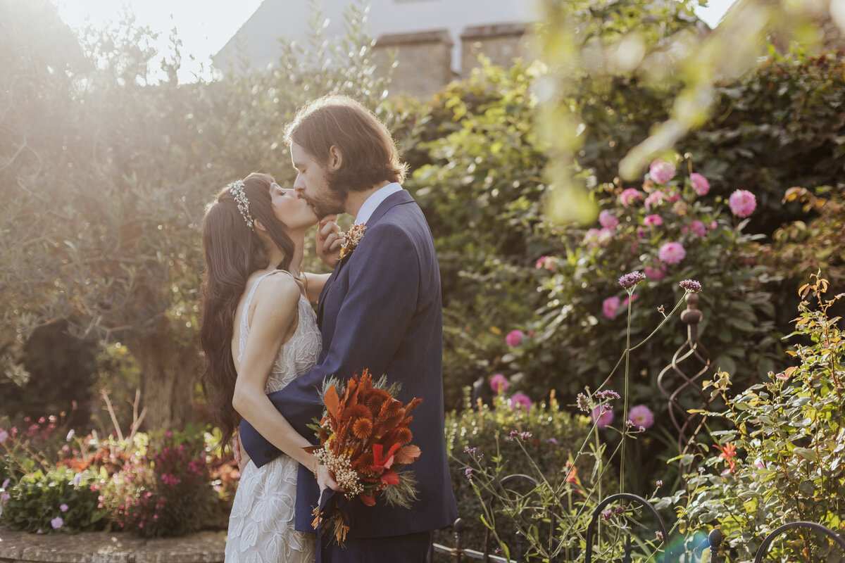 Bride and groom kiss in dreamy golden hour light, in a garden filled with flowers