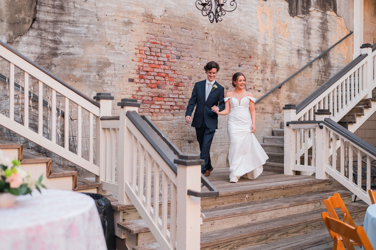 A bride in a white dress and a groom in a dark suit walk arm-in-arm down wooden stairs outside, smiling, with a weathered brick wall in the background.