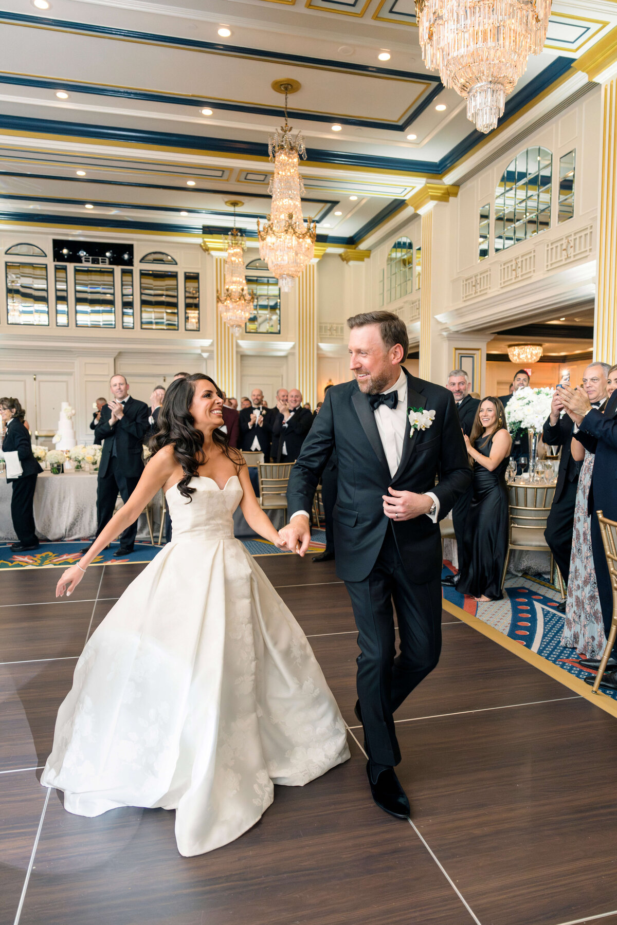 A bride and groom joyfully dance at their wedding reception. The bride wears a strapless white gown and the groom a black tuxedo. Guests in formal attire applaud around them in an elegant, chandelier-lit ballroom.