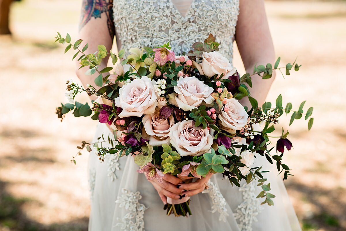 Detail photo of a bridal bouquet. The bride is wearing a silver lace wedding dress and she is holding a horizontal cascade bouquet of  blush and light pink roses and burgundy accent flowers with greenery and eucalyptus.