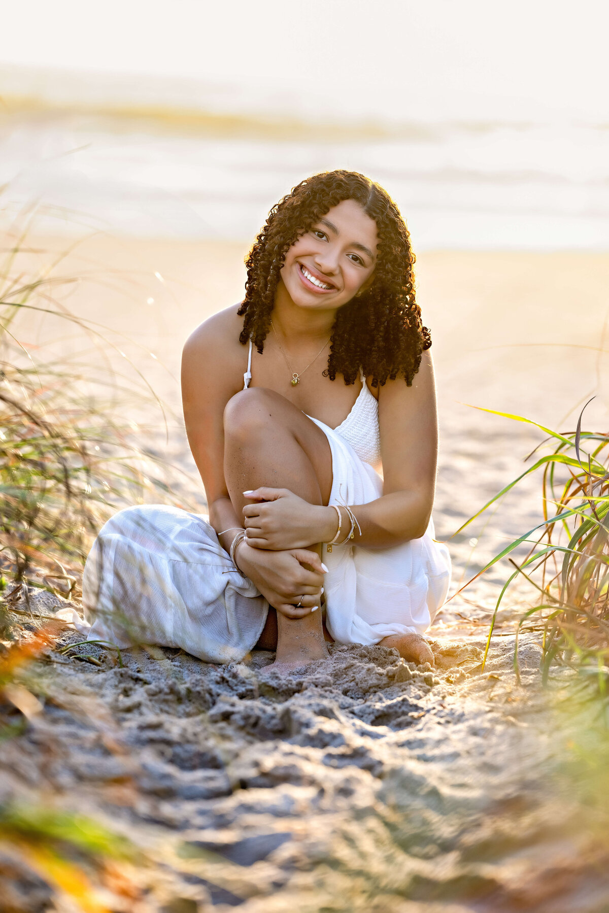 Winter Garden senior photos in beach grass with a teen girl smiling, legs up, wearing a white sundress.