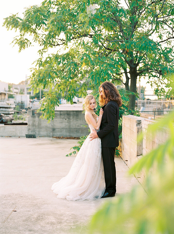 Bride and groom wearing a black tuxedo and white wedding gown holding each other outside on a rooftop
