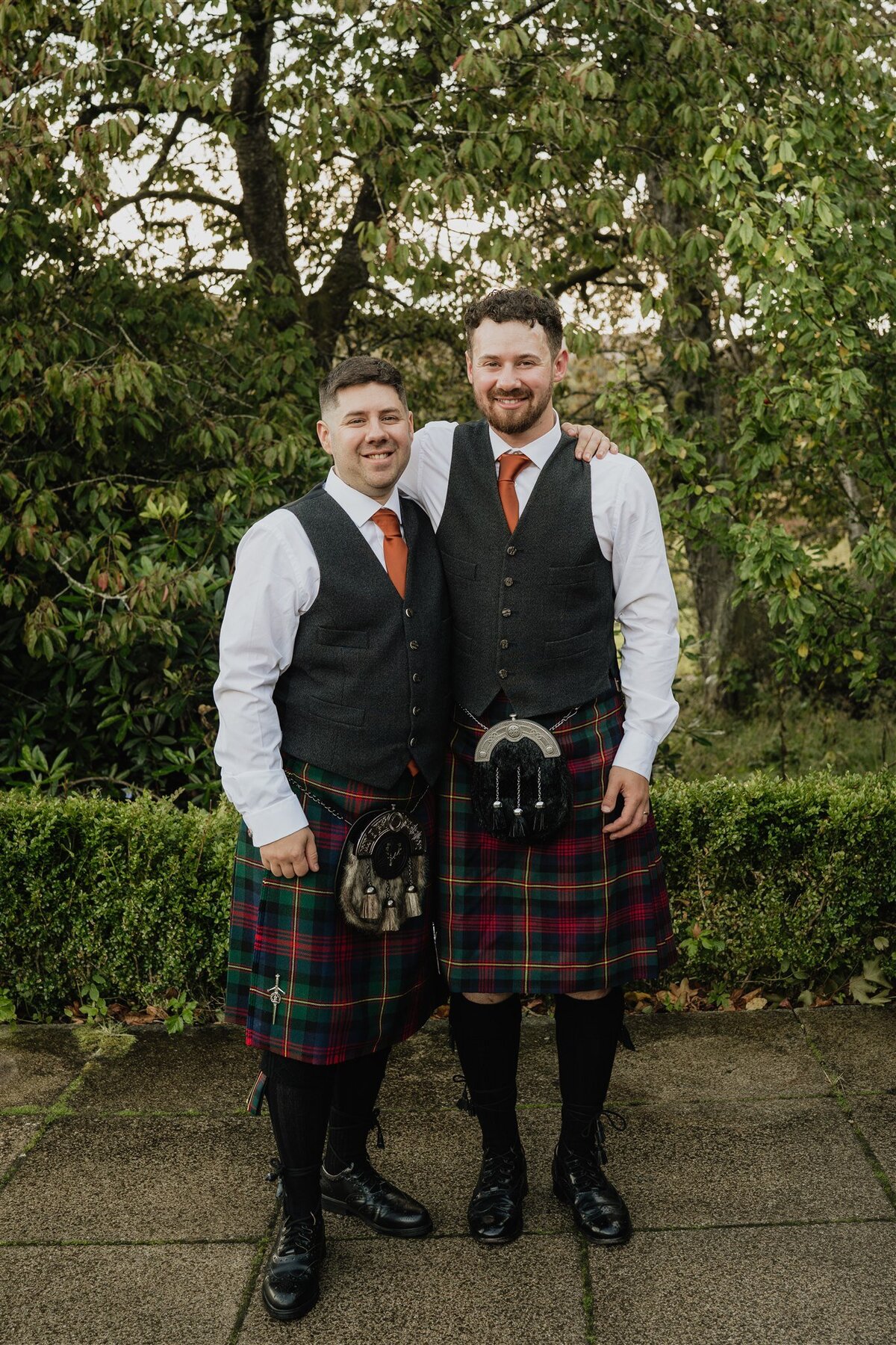 Two men stand side by side and pose for a photograph. They both wear kilts and traditional Scottish wedding attire. Trees and greenery surround them.