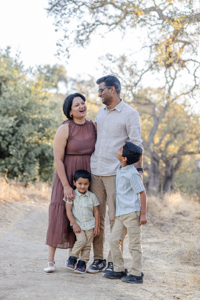 Family of four pose happily looking at each other during a golden hour fall photoshoot at an Oak Grove in San Jose