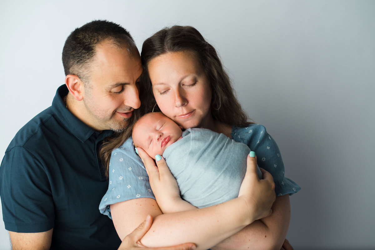 Family portrait with mom and dad standing close, wearing blue color outfits, mom is holding newborn baby white they snuggle in close with eyes closed.