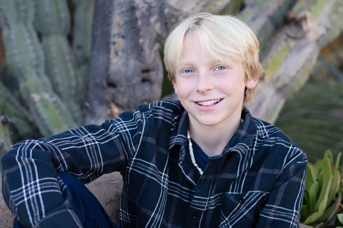 A close-up of a blonde teenager in a dark blue and white striped flannel, smiling up at the camera in San Juan Capistrano.