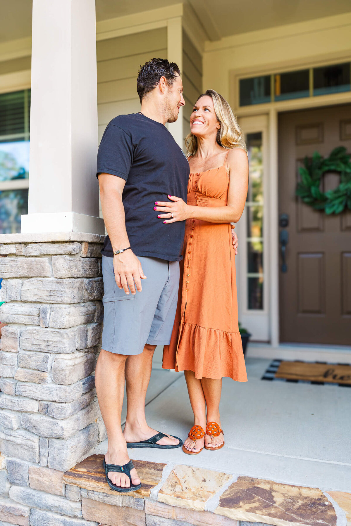 A couple stands on a porch, framed by the lens of a family photographer in Fayetteville, NC. The man dons a black shirt and gray shorts, while the woman dazzles in an orange dress and sandals. They smile warmly at each other beside an entrance door adorned with a green wreath.