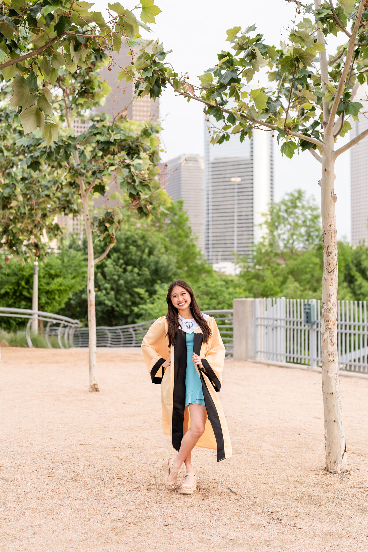 Houston High School senior girl wearing gown and blue dress with hand on hip and holding gown while standing in middle of park with Downtown Houston in the background
