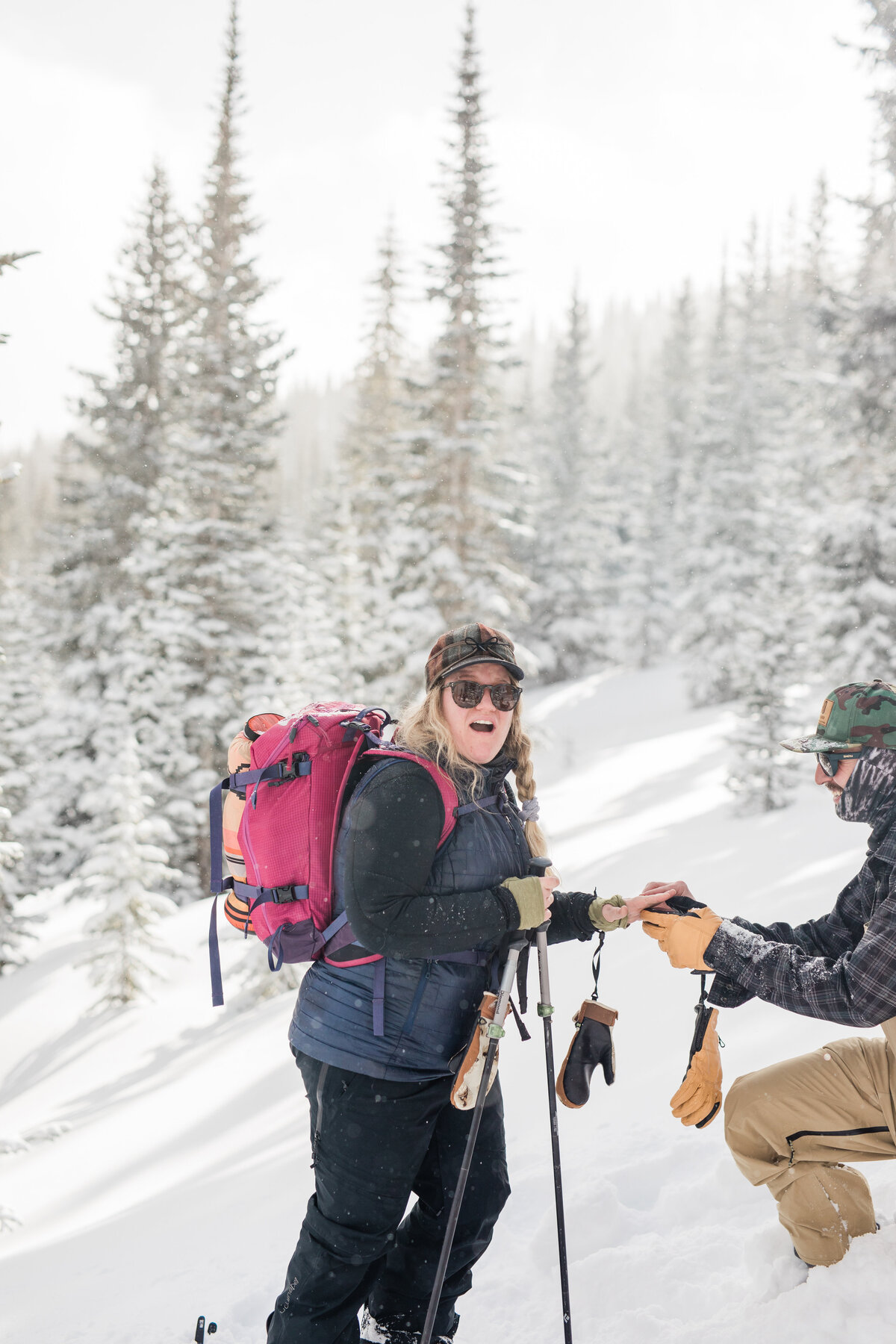 A woman in all backcountry gear looks surprised at the camera as her boyfriend gets down on one knee and she realizes she is being proposed to