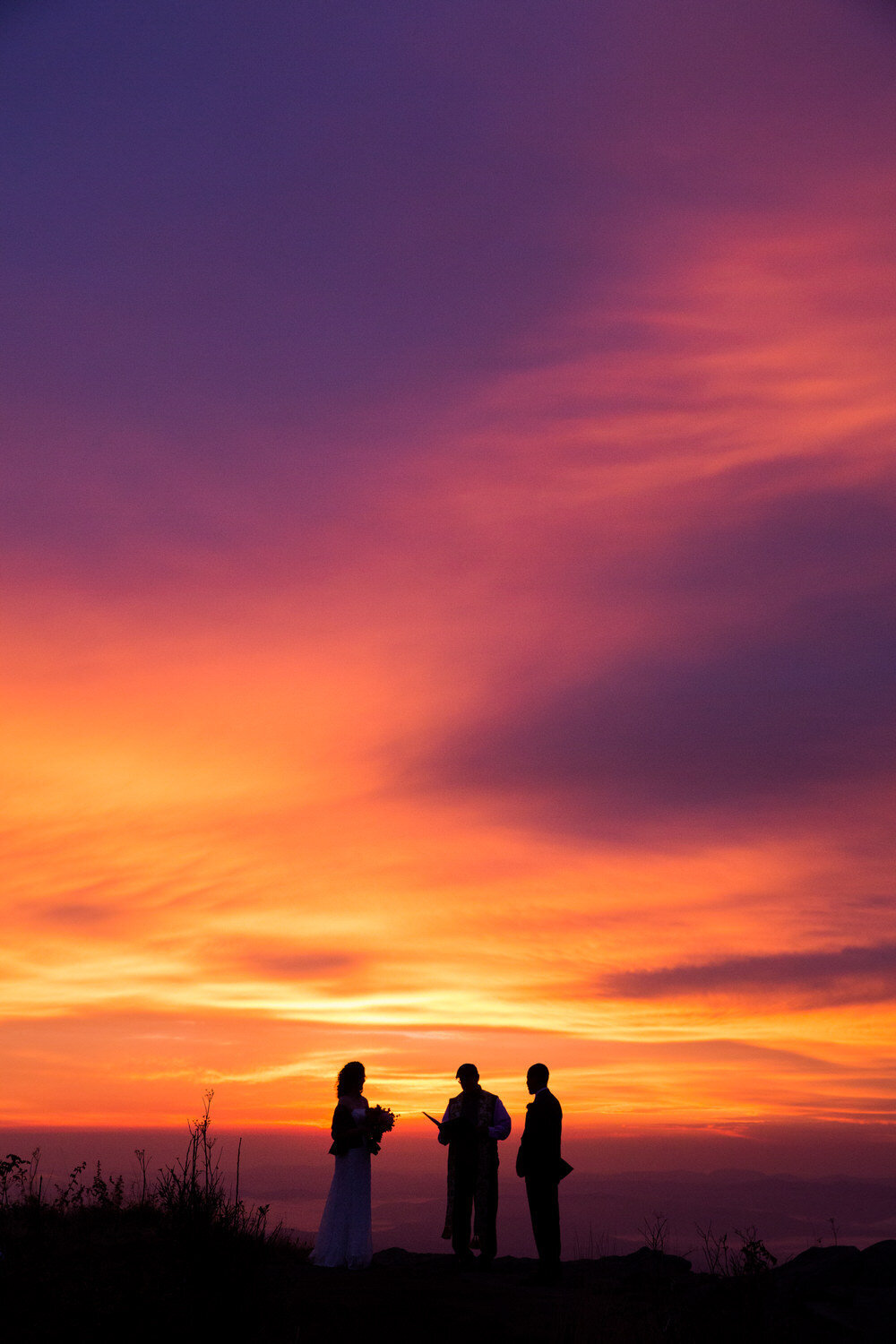 couple stands against an epic colorful sunrise at their elopement ceremony with their officiant