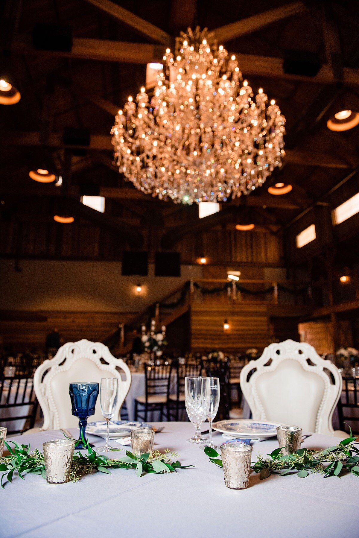 The bride and groom's reception table at Sycamore Farms is set with blue mismatched vintage china, cobalt blue water goblets, gold votive candles and garlands of greenery. The bride and groom have large elaborately decorated carved ivory throne chairs set at their table.