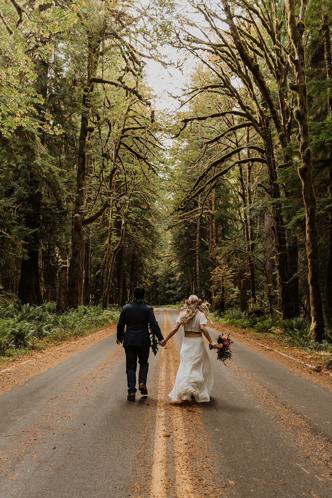 Bride & groom holding hands while running down road through old growth forest in Olympic National Park!
