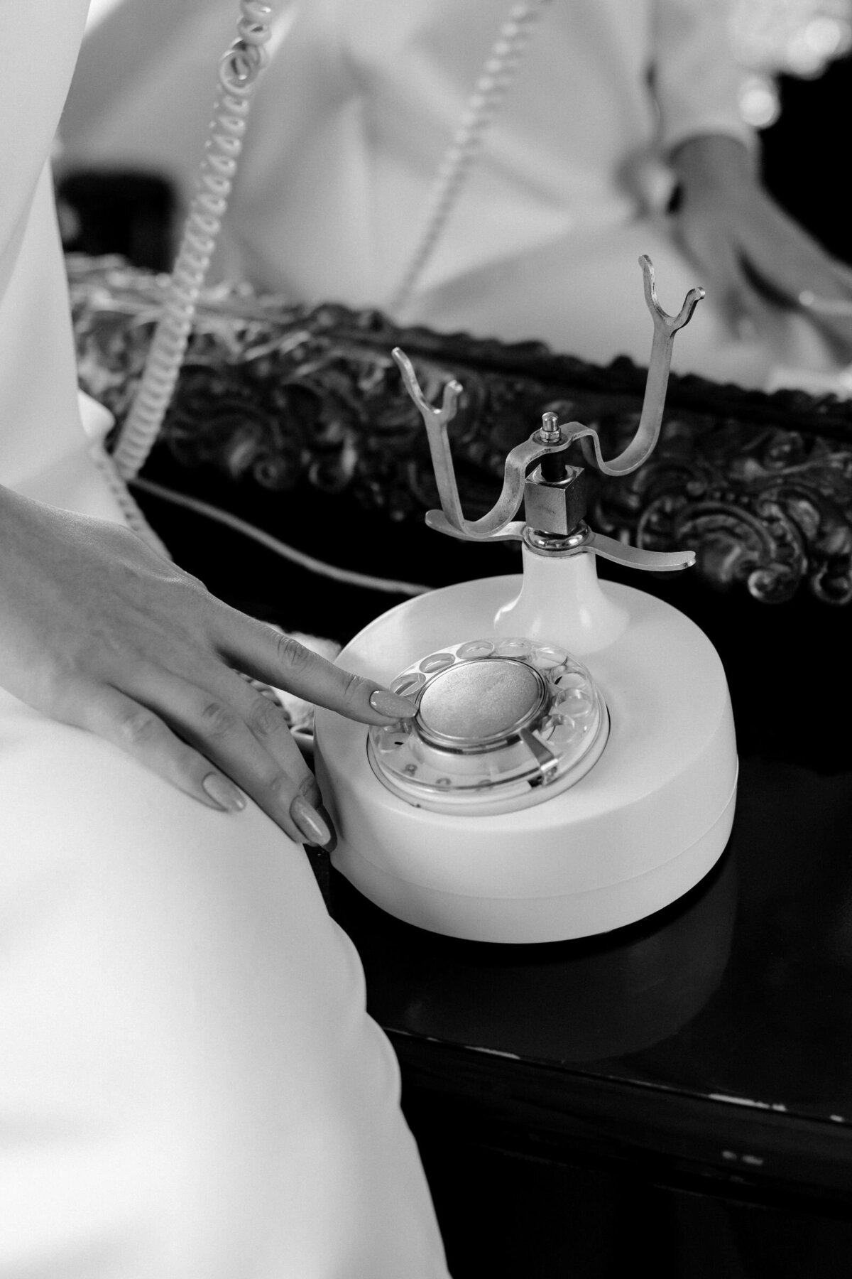 Close-up of a woman with her fingers poised to dial a number on a vintage rotary phone. She wears an elegant white silk dress, sitting atop a black dresser with an ornate framed mirror in the background, capturing a moment of refined vintage charm.