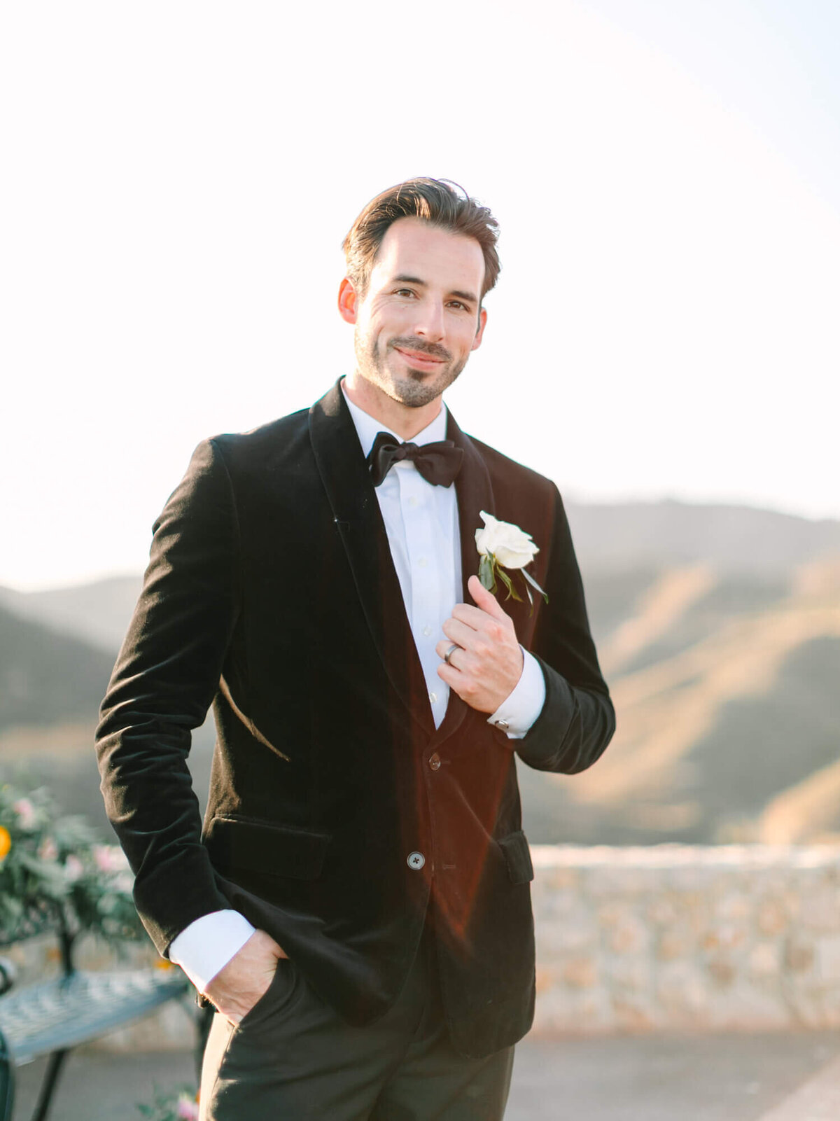 Groom stands smiling in a black tuxedo with a white boutonnière, hands in pockets, against a sunlit mountainous backdrop.