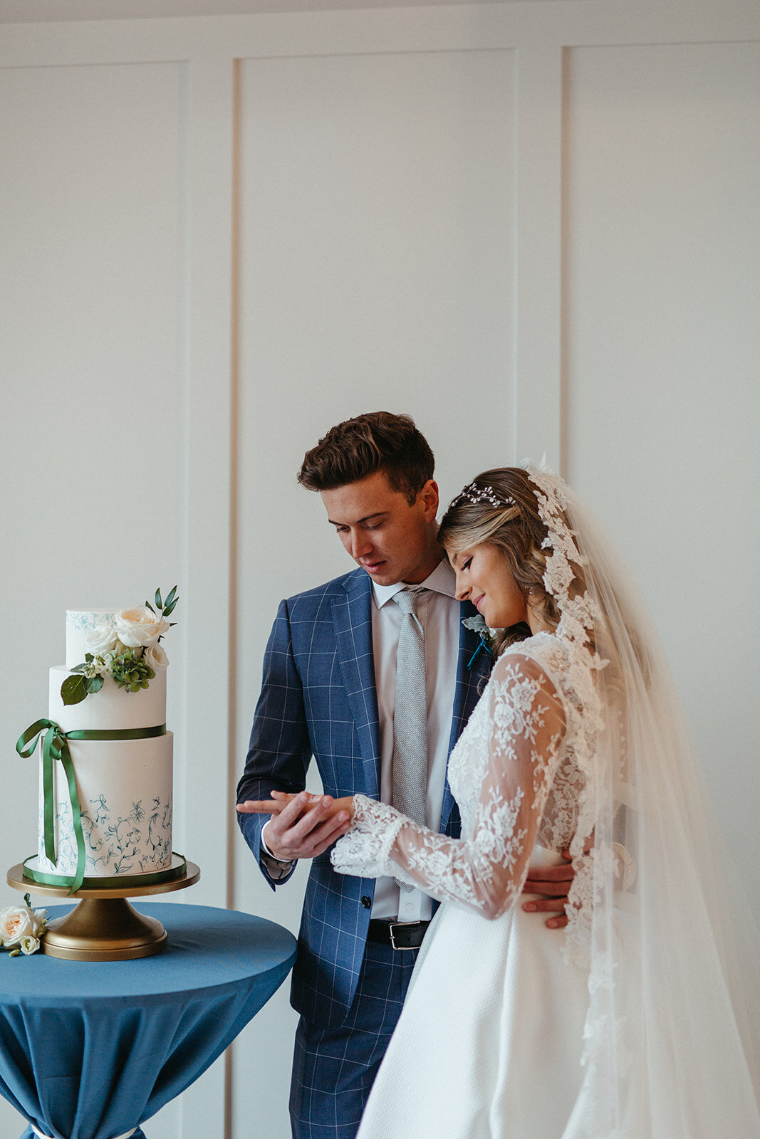 Bride and groom in a blue suit and white wedding gown next to a layered cake on gold cake stand atop a round table.