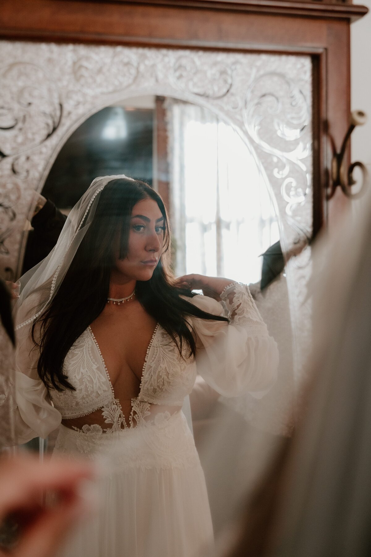 Bride in lace wedding dress adjusting her veil while looking in an ornate mirror, captured in soft natural light.