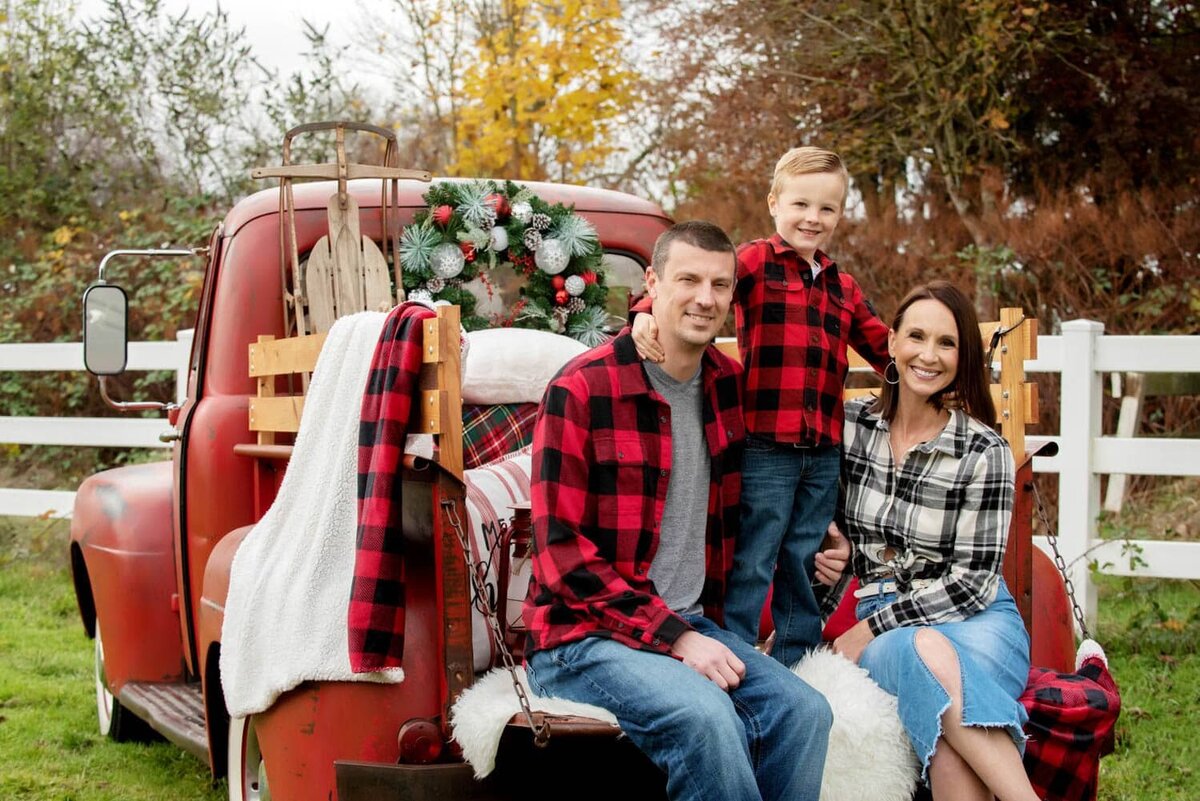 Cute holiday plaid family, mom and dad sitting with standing son in between on vintage red truck decorated for Christmas.