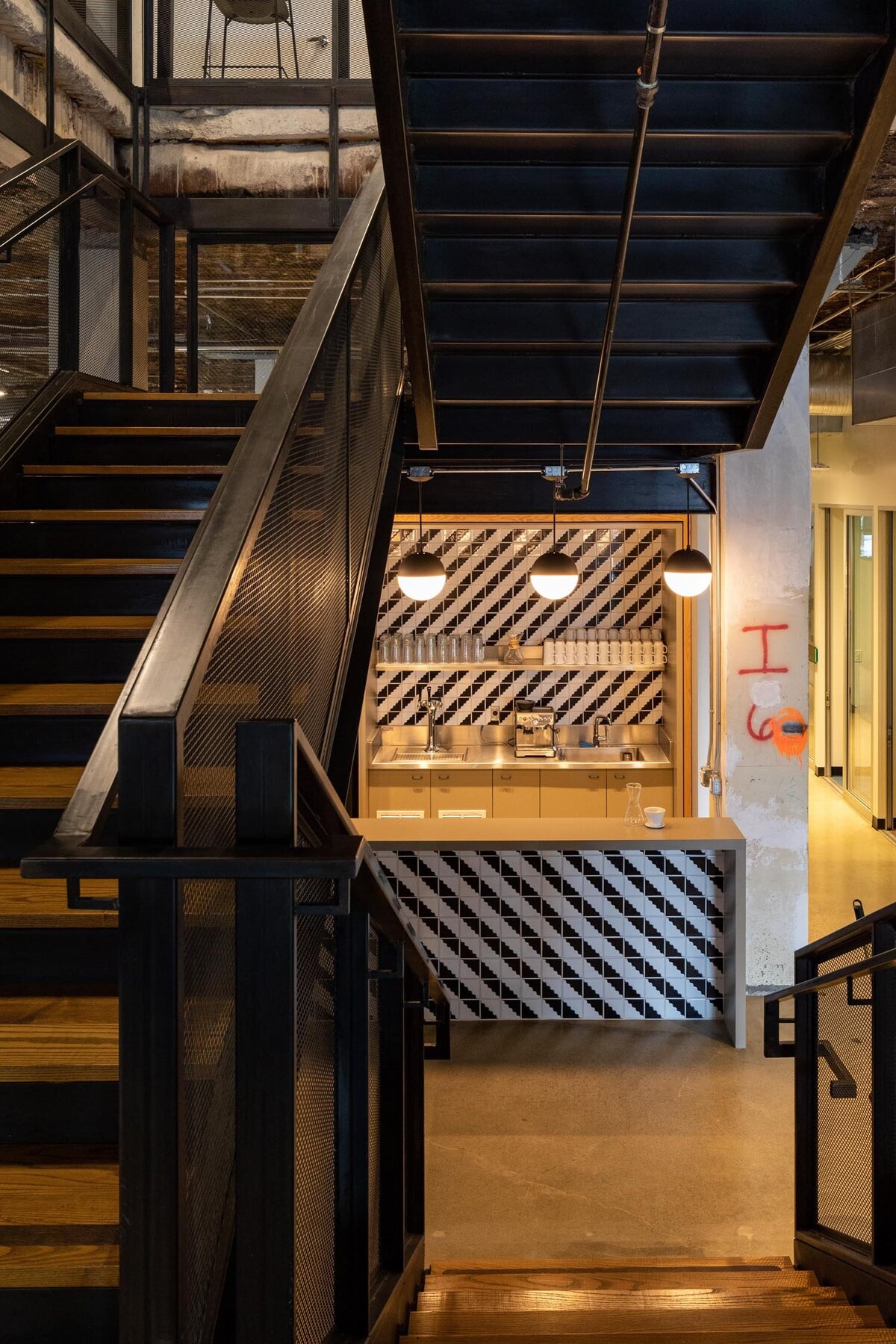 Industrial staircase leading to a small coffee station with geometric black-and-white tile and globe pendant lighting.