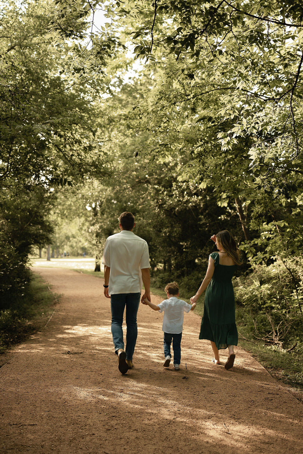 A family of three walks hand in hand down a tree-lined dirt path. Sunlight filters through the leaves, creating a peaceful, warm atmosphere.
