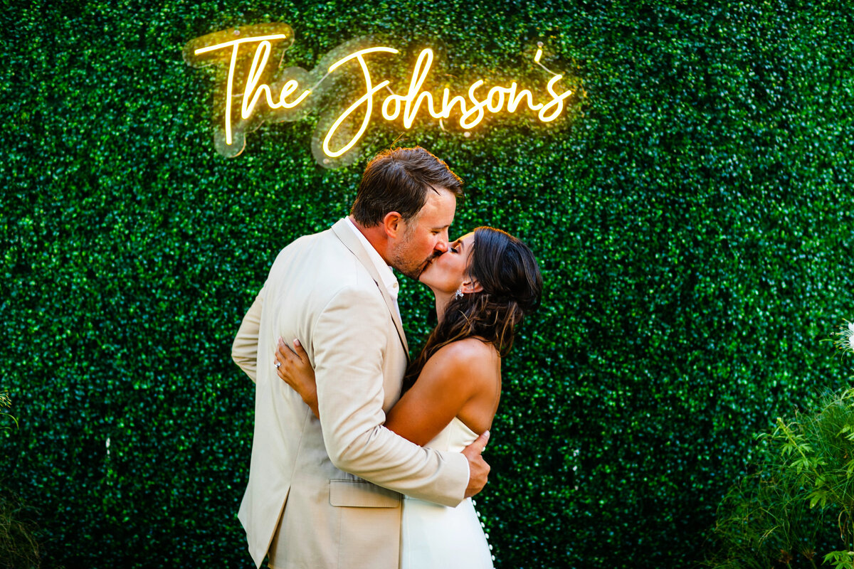 A bride and groom kiss under a neon sign with their new last name.