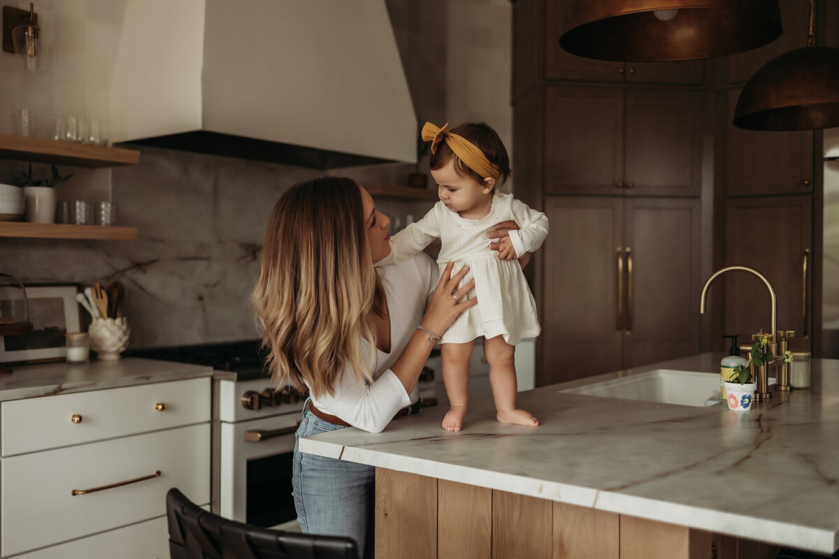 Mom and toddler girl embrace in kitchen while toddler stands on kitchen island